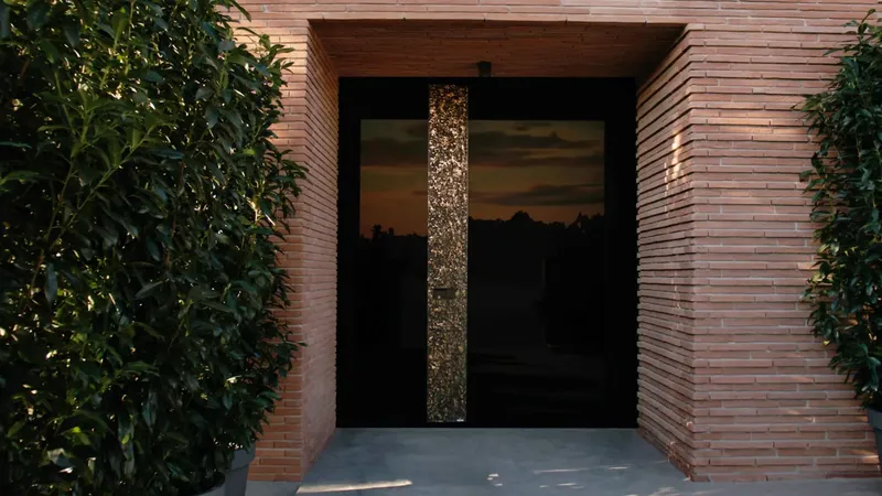 Črna vrata z zlatim okrasnim stebrom so obkrožena z opečnatimi stenami in zelenjem.