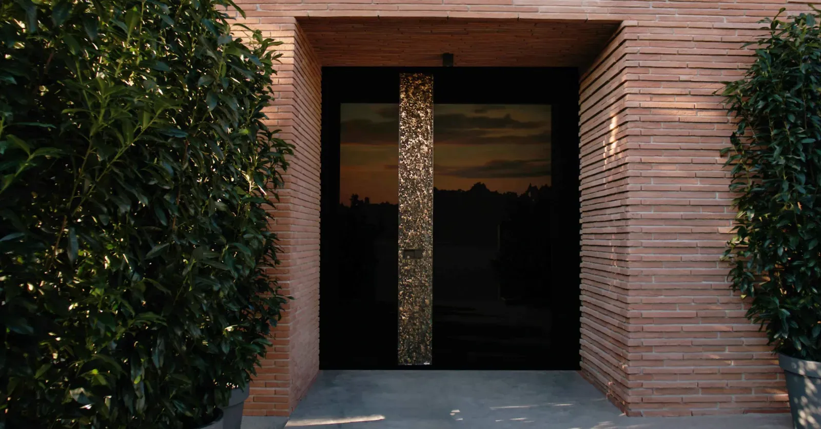 Črna vrata z zlatim okrasnim stebrom so obkrožena z opečnatimi stenami in zelenjem.