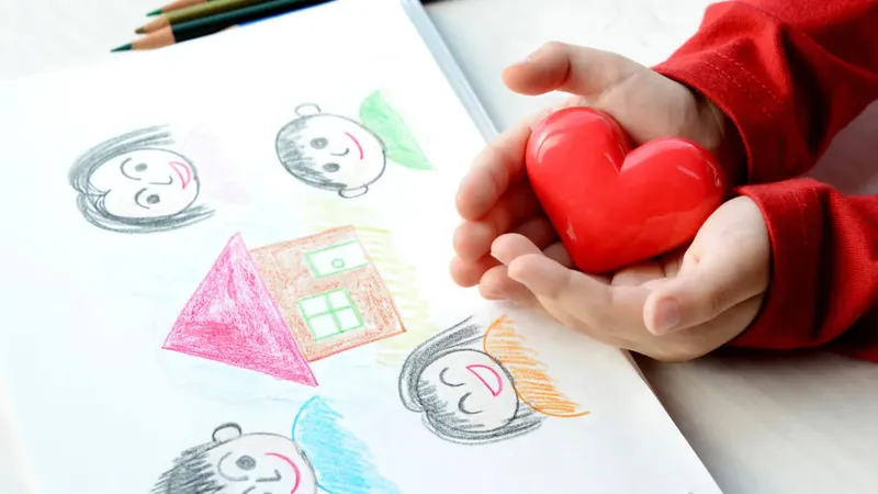 Otrok drži rdeče srce v dlaneh ob risbi družine in hiše, narisani s pisanimi barvicami.