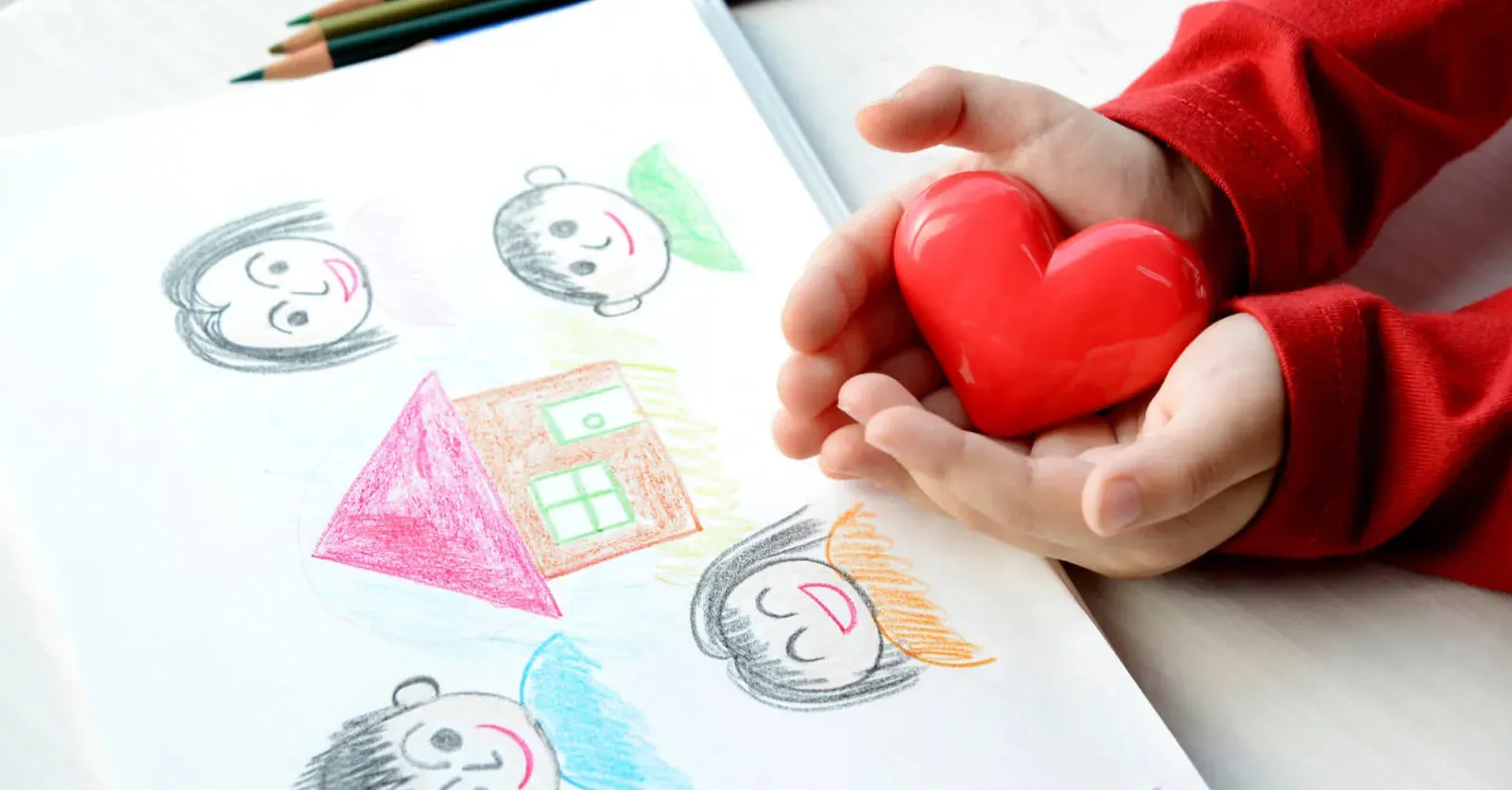 Otrok drži rdeče srce v dlaneh ob risbi družine in hiše, narisani s pisanimi barvicami.