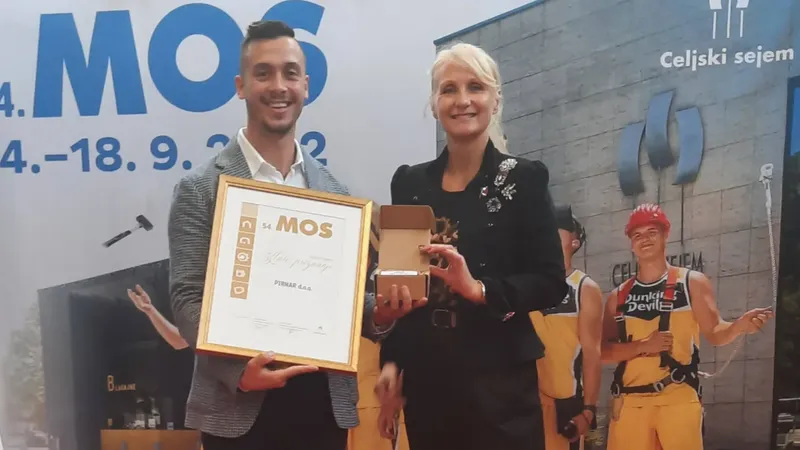 Moški in ženska držita priznanje in darilo na odru, v ozadju so ljudje v delovnih oblačilih.