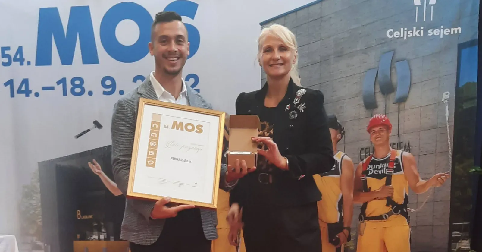 Moški in ženska držita priznanje in darilo na odru, v ozadju so ljudje v delovnih oblačilih.