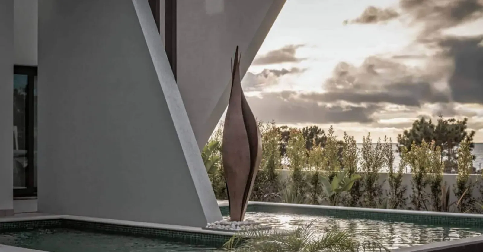 Pergola iz betona s kovinsko skulpturo ob bazenu in zelenjem v ozadju.