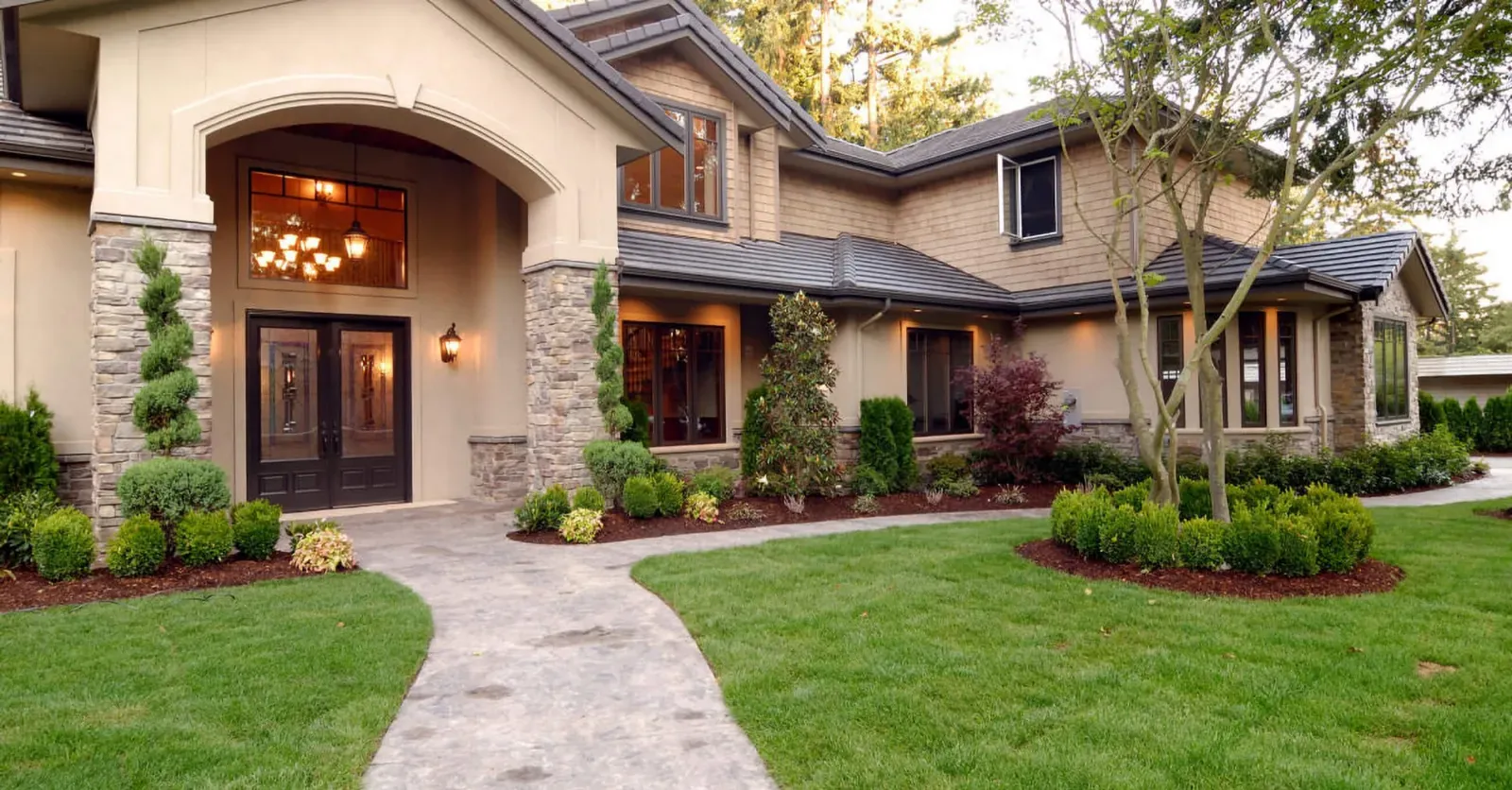 Dve leseni vhodni vrata z okni in svetilkami, obkrožena z zelenjem in tlakovanjem.