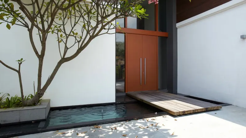 Vhodna vrata so rjava z dvema kovinskima ročajema, obdanimi z belimi stenami in drevesom.