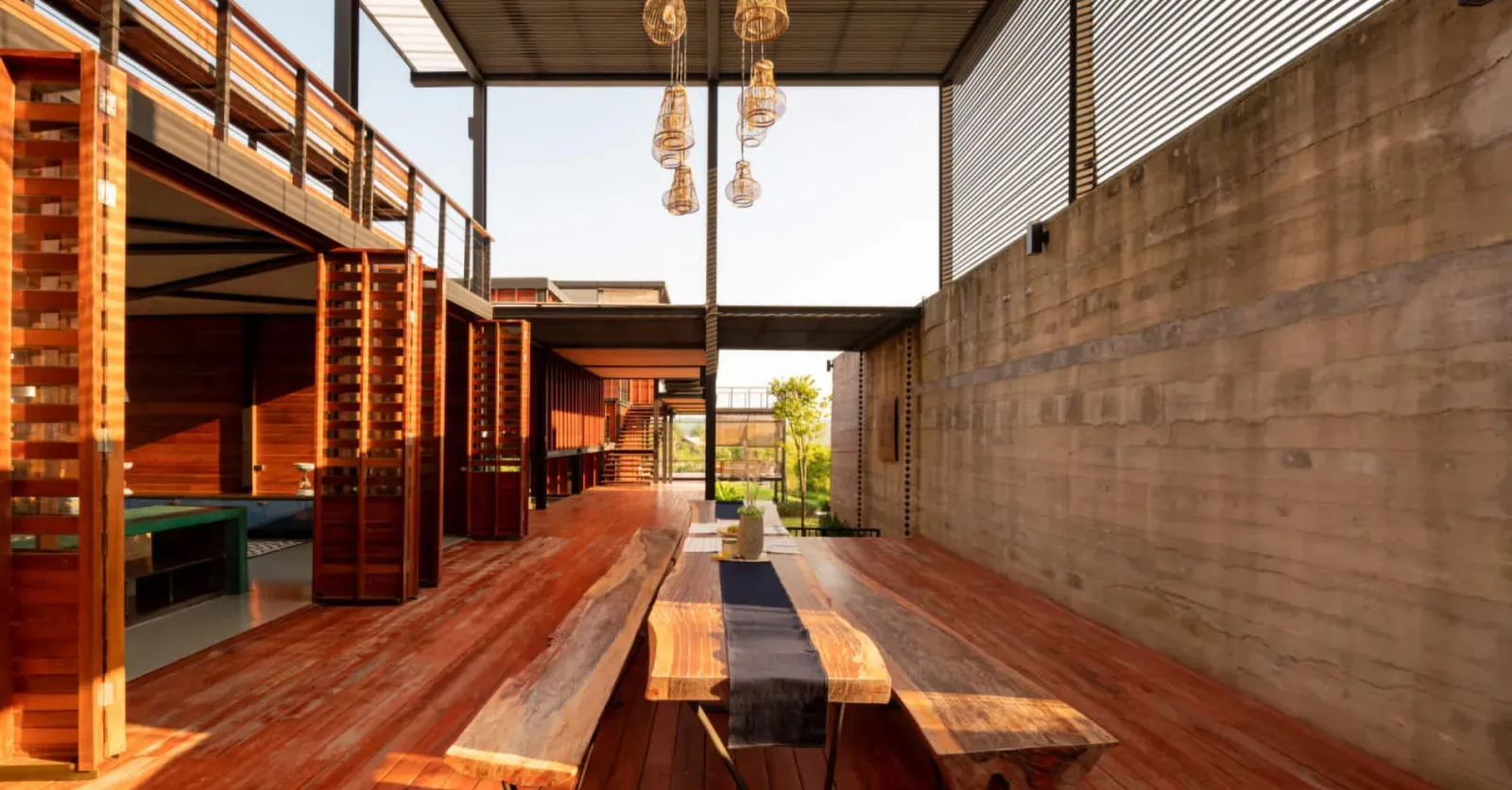 Lesena pergola s visečimi svetili in dolga lesena miza s prtom v središču prostora.