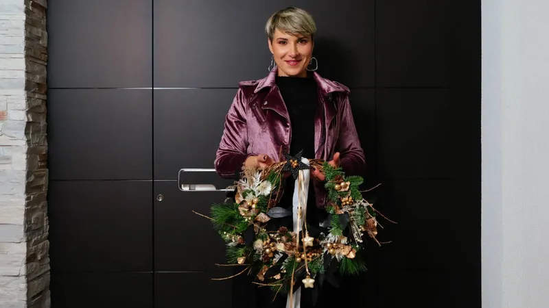 Črna vhodna vrata z ravnimi paneli in oseba, ki drži venec iz zelenja in dekorativnih elementov.