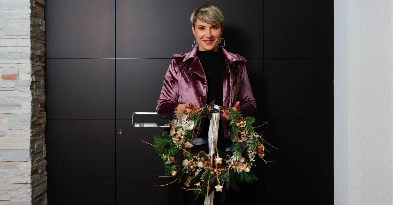 Črna vhodna vrata z ravnimi paneli in oseba, ki drži venec iz zelenja in dekorativnih elementov.