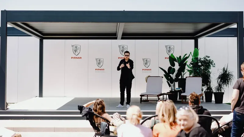 Pergola z ravnim streho in odprtim prostorom, obdan z rastlinami in sedežno garnituro.