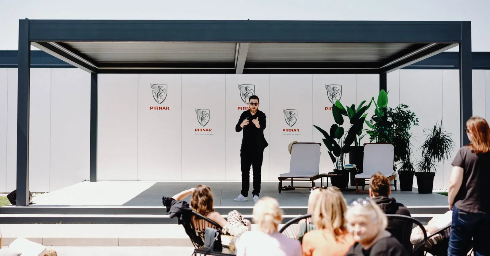 Pergola z ravnim streho in odprtim prostorom, obdan z rastlinami in sedežno garnituro.