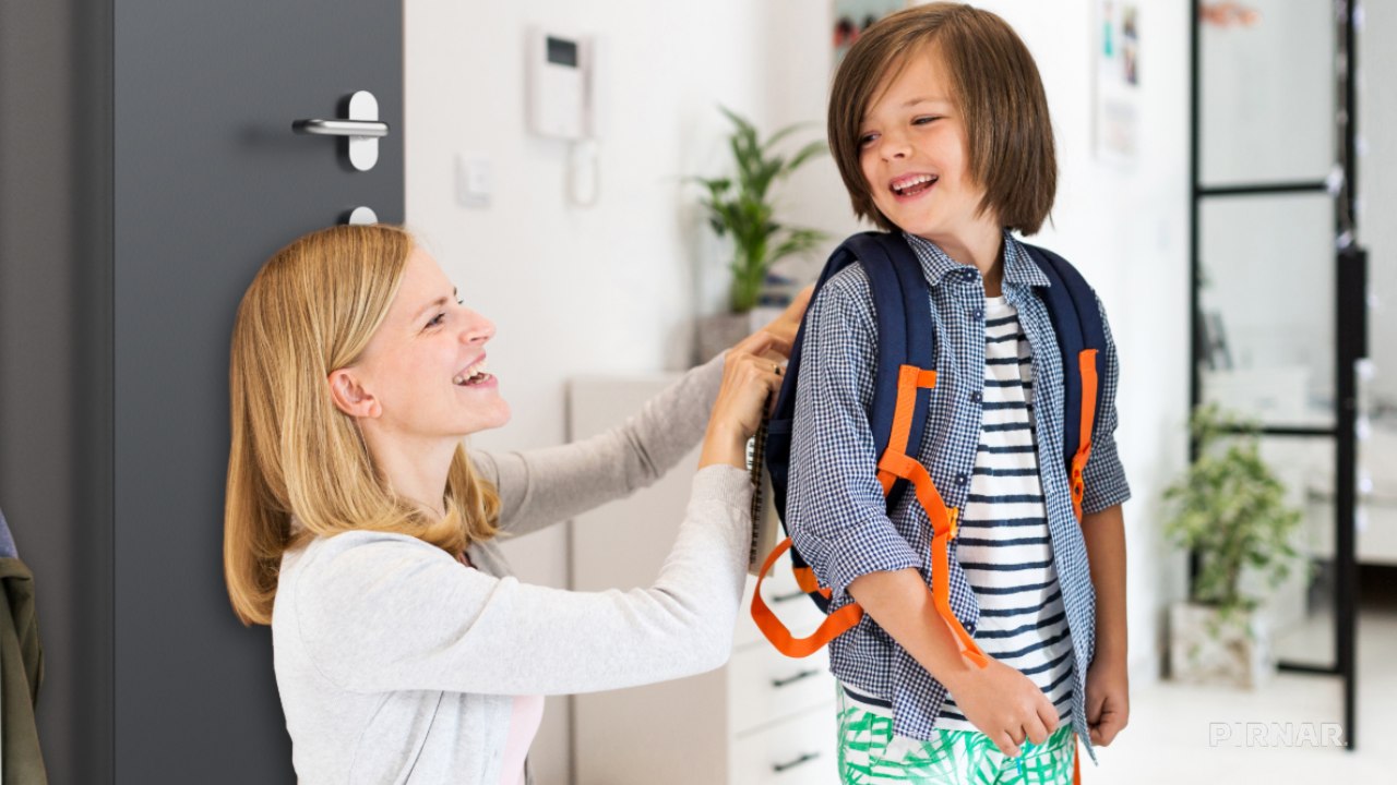 Ženska pomaga otroku z nahrbtnikom pri vhodnih vratih, oba se nasmejana pripravljata na odhod od doma.