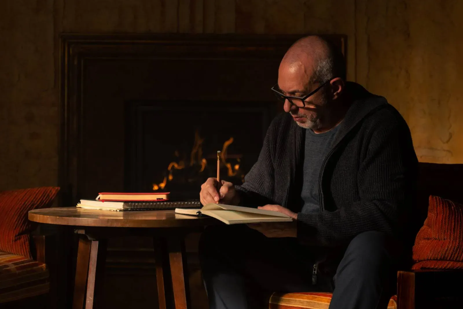 Moški piše v zvezek, medtem ko sedi za leseno mizo ob ognju.