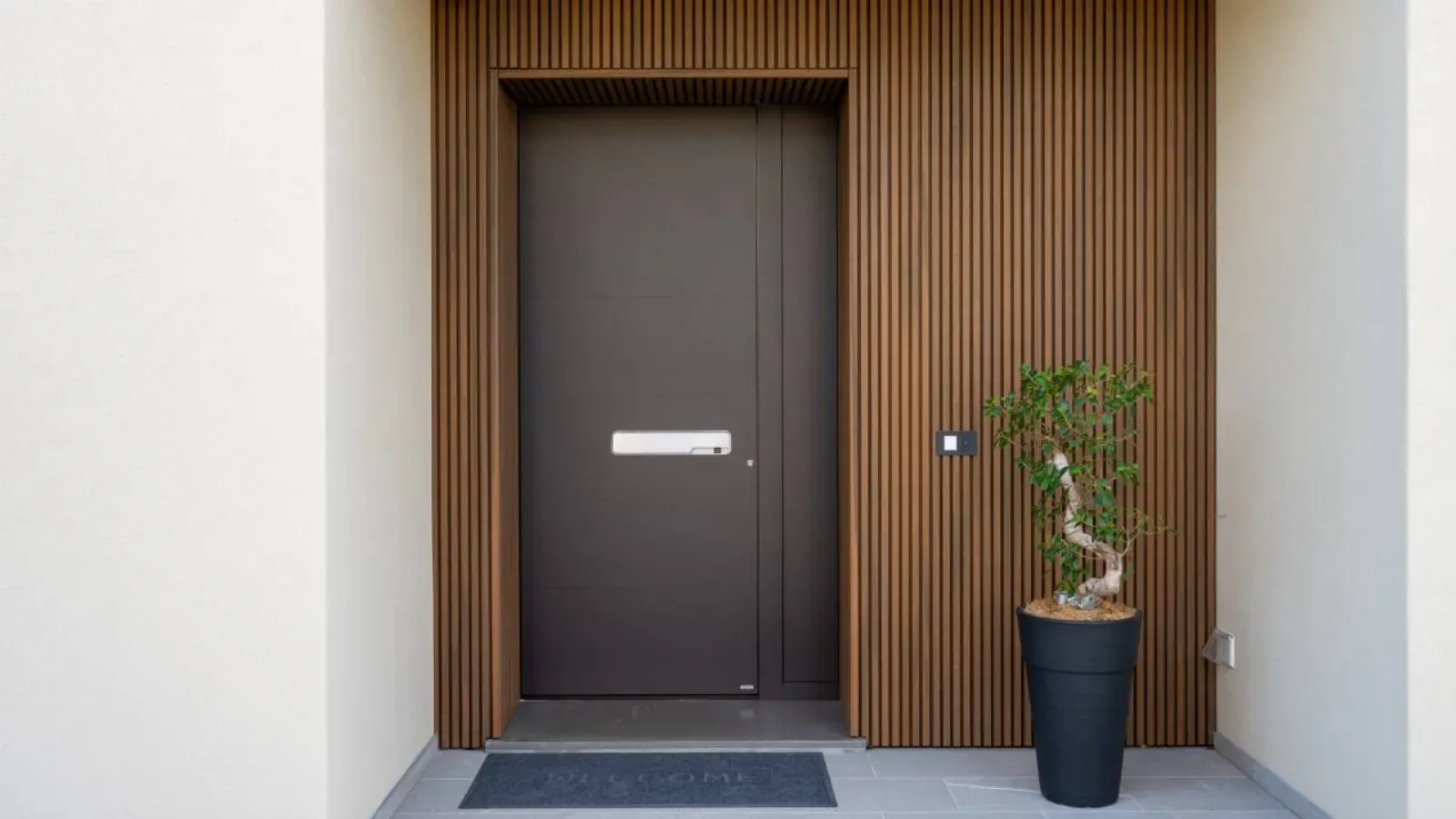 Vrata s preprosto obliko in kovinsko ročico so obkrožena z lesenimi letvami, ob strani pa je lončnica z rastlino.